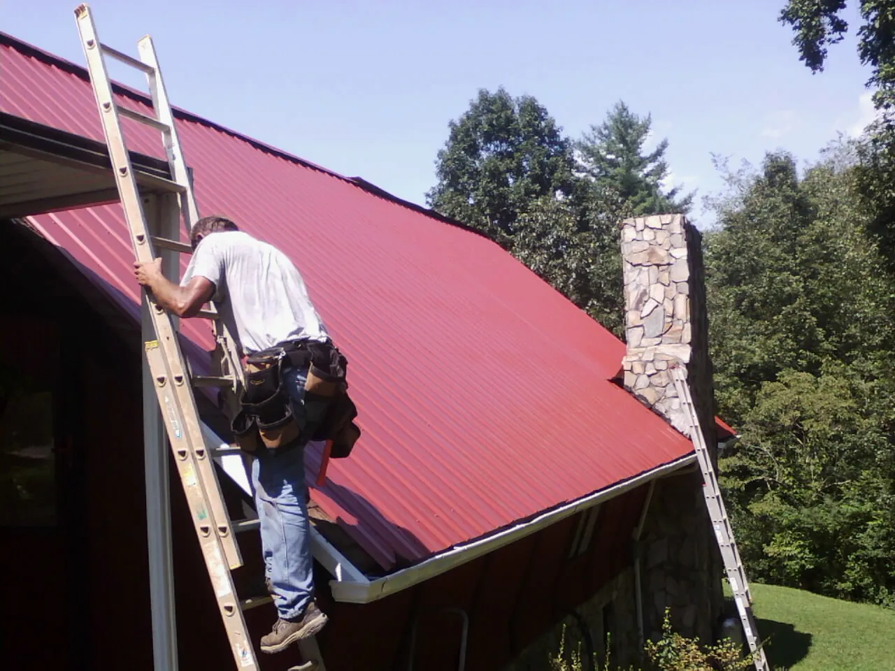 Roofer on a ladder beside a red metal roof and stone chimney — Soffit & Fascia Repair by Roofing Brothers in Mayfield